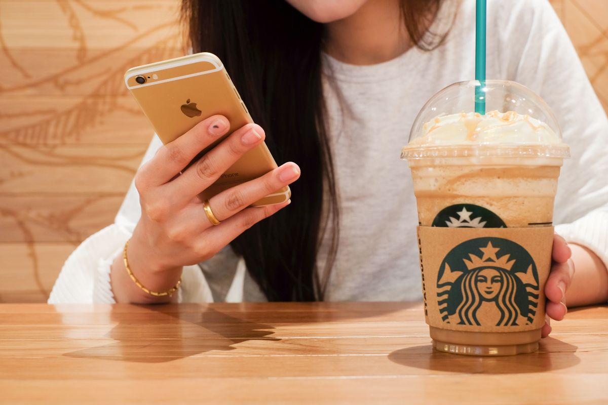 A woman with long dark hair sits ata table holding an iphone and a starbucks caramel frappuccino