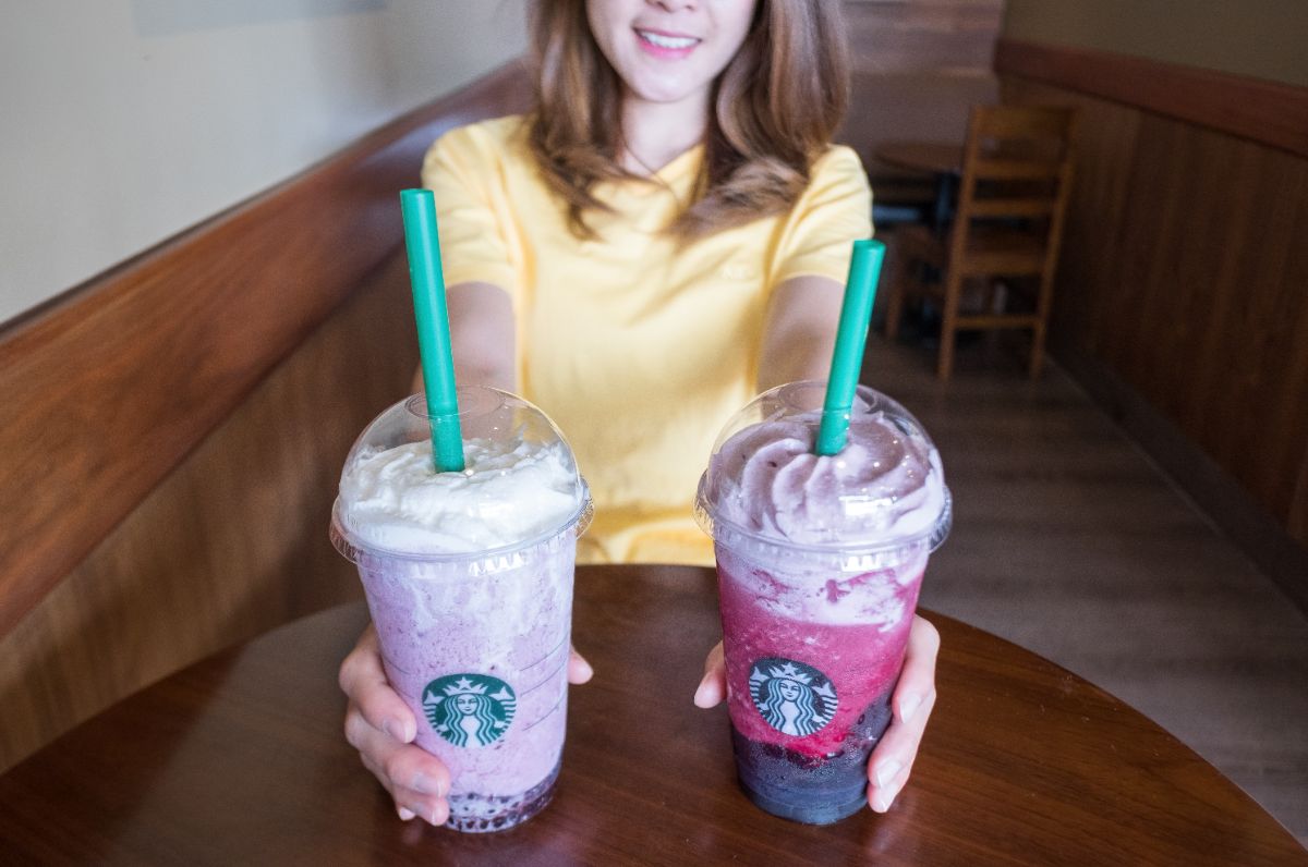 a woman in a yellow shirt holds out two starbucks Refreshers, one purple and one violet